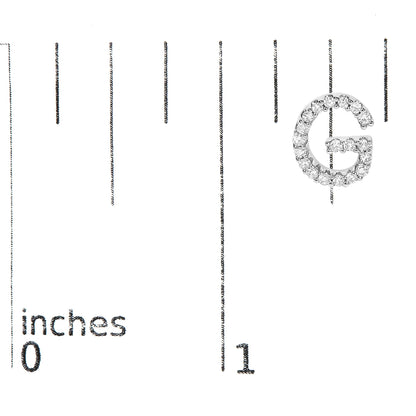Close-up top-down view of a white metal earring shaped like the letter G, covered in small white gemstones, shown on a scale marked in inches.