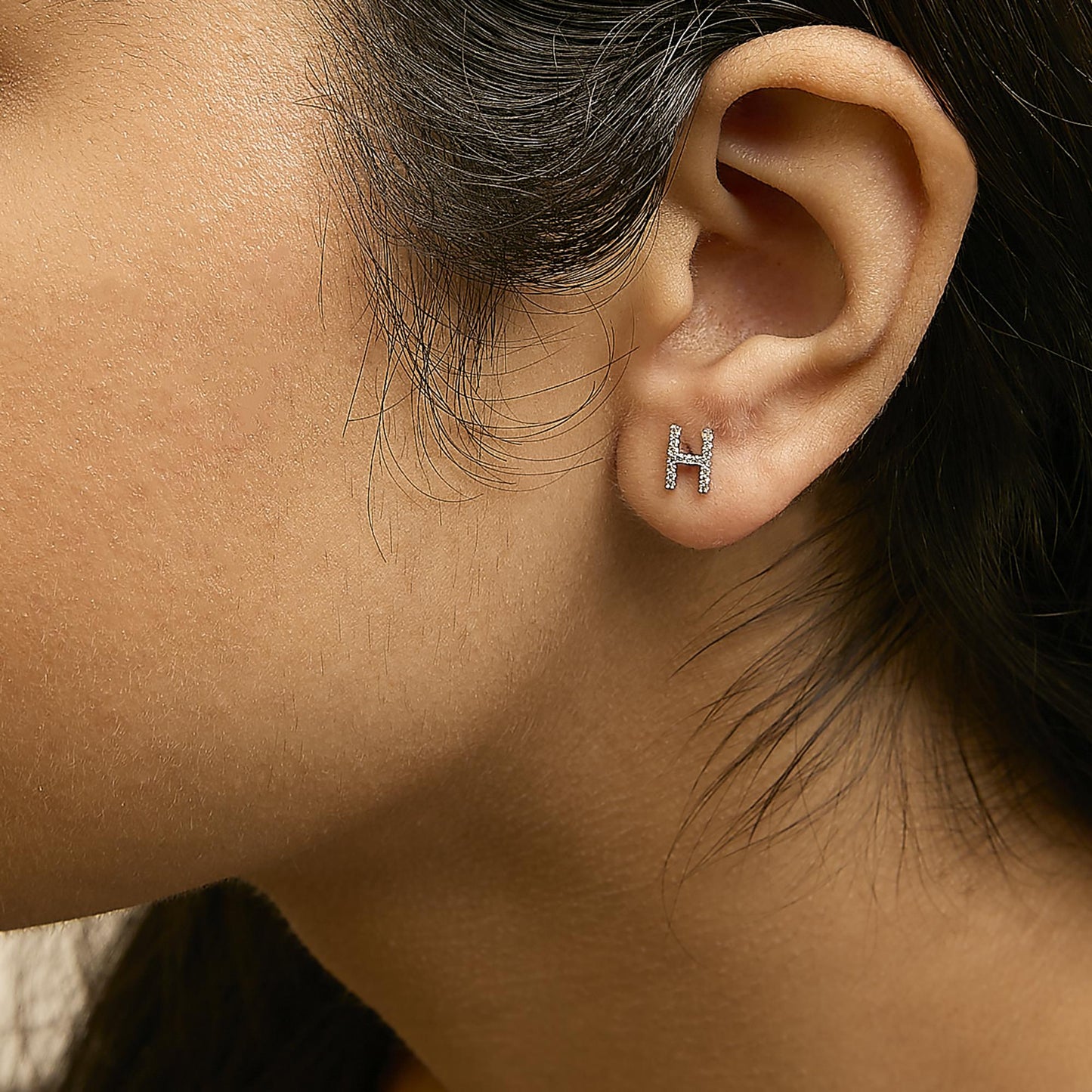 Close-up side view of a model's ear wearing a white metal earring shaped like the letter H, adorned with multiple small white gemstones in a micro-pave setting.