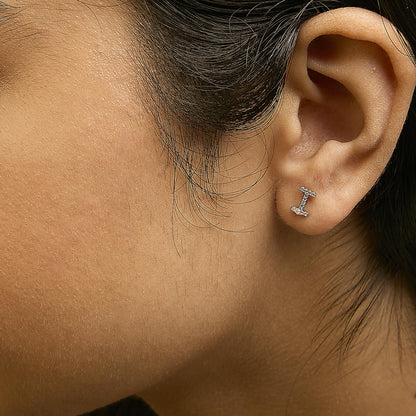 Close-up side view of a model wearing a white metal earring shaped like a horizontal bar with two vertical ends, featuring small white gemstones in a micro-pave setting.