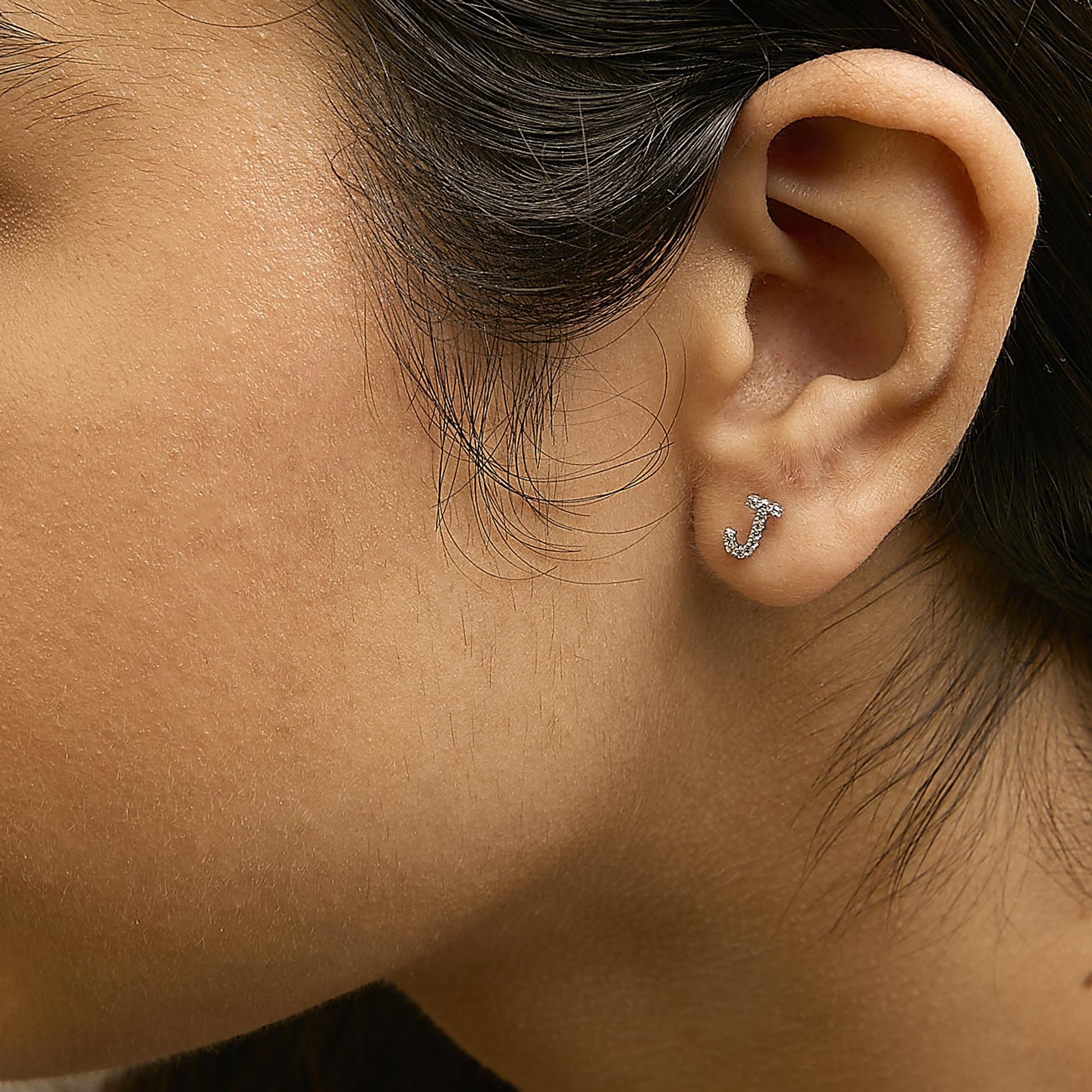 Close-up side view of a model wearing a white metal earring shaped like the letter 'J' encrusted with small round white gemstones in a micro-pave setting.