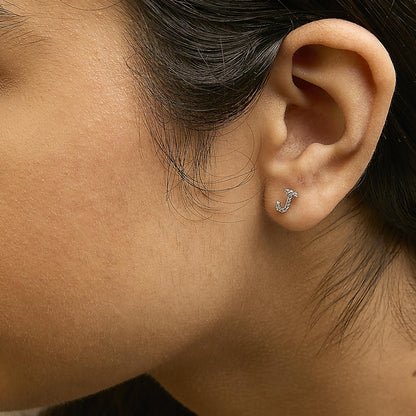 Close-up side view of a model wearing a white metal earring shaped like the letter 'J' encrusted with small round white gemstones in a micro-pave setting.