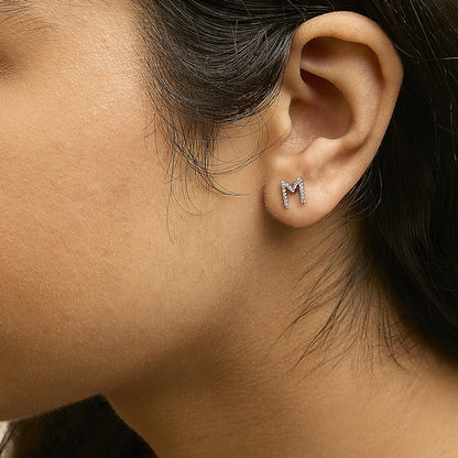 Close-up side view of a model wearing white metal stud earrings shaped like the letter M, adorned with round white gemstones in a micro-pave setting.