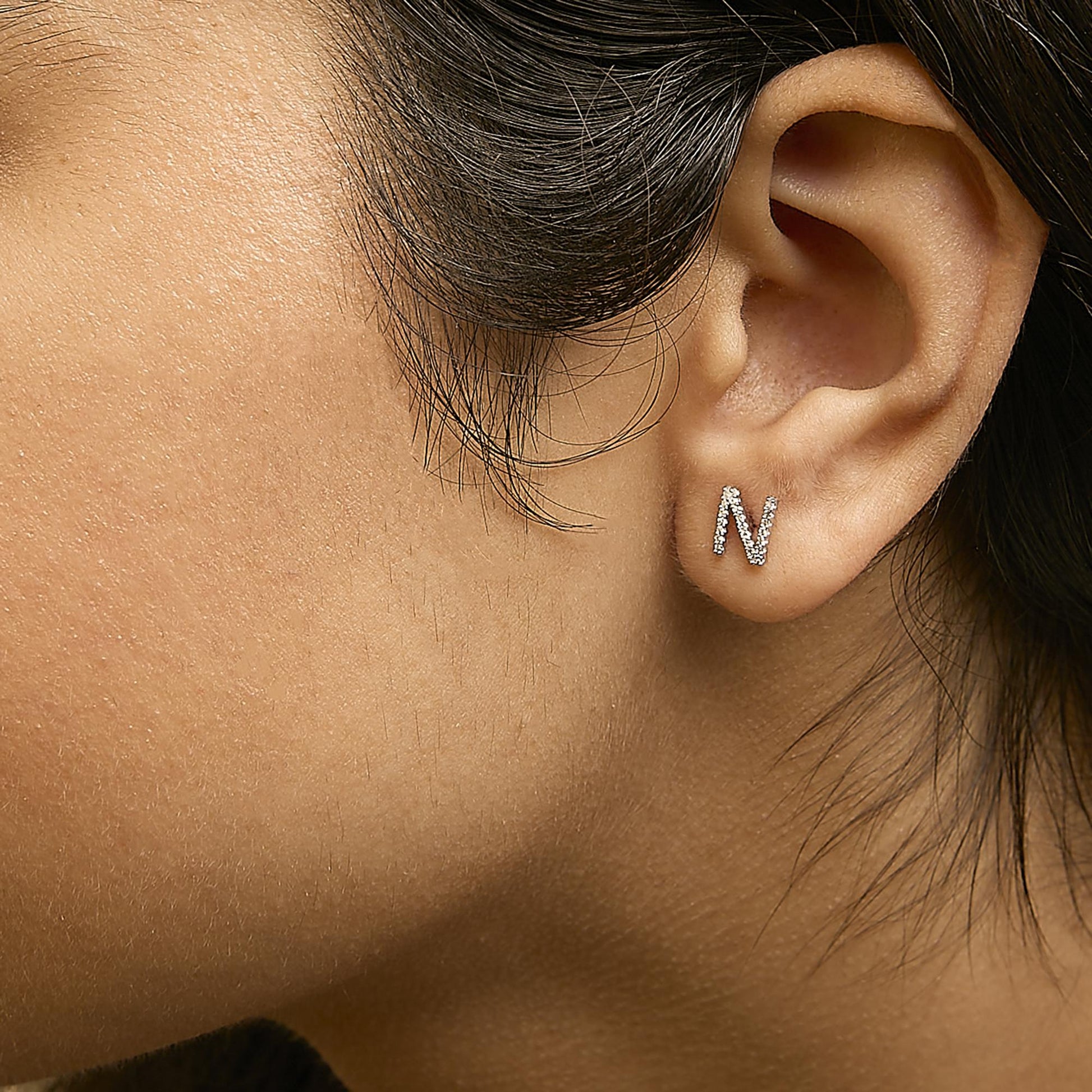 Close-up side view of a model wearing a white metal earring shaped like the letter N, adorned with small round white gemstones in a micro-pave setting.