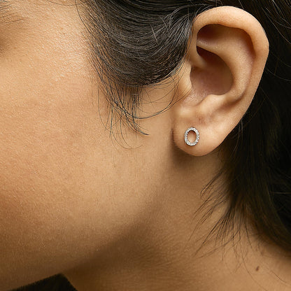 Close-up side view of a model wearing white metal oval-shaped earrings with round white gemstones set in a micro-pave style.