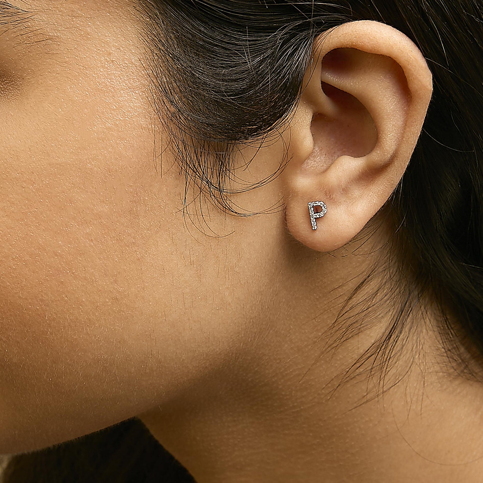 Close-up side view of a model wearing white metal earrings shaped like the letter P, featuring round white gemstones in micro-pave setting.