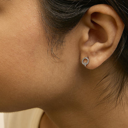Close-up side view of a model wearing small white metal earrings shaped like a loop with round white gemstones in a micro-pave setting.
