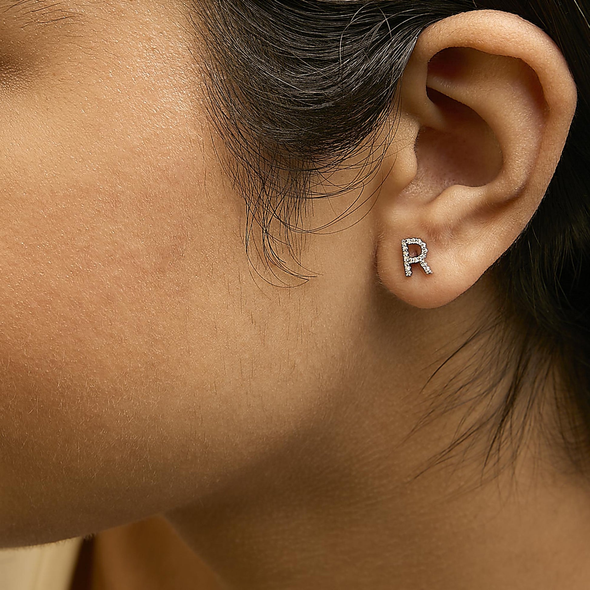 Close-up side view of a white metal earring shaped like the letter R, adorned with small white gemstones, shown worn on a model's ear.