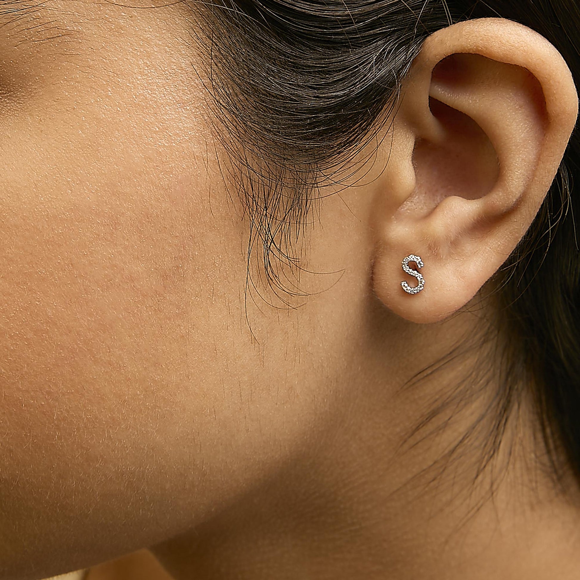 Close-up side view of a white metal letter 'S' shaped earring with round white gemstones in micro-pave setting, shown worn on a model.