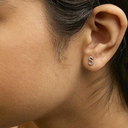 Close-up side view of a white metal letter 'S' shaped earring with round white gemstones in micro-pave setting, shown worn on a model.
