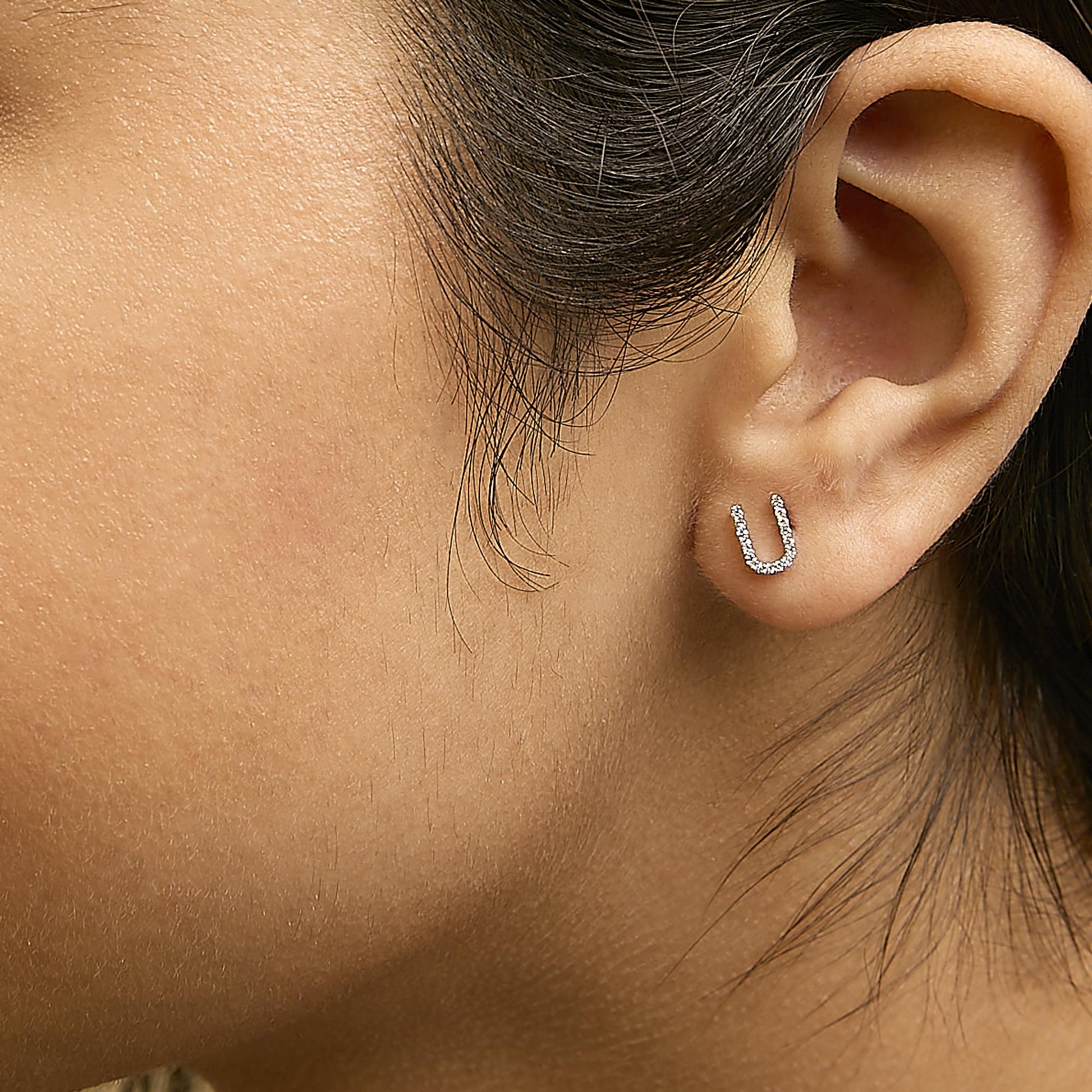 Close-up side view of a model wearing white metal earrings shaped like a small horseshoe, featuring micro-pave set white round gemstones.