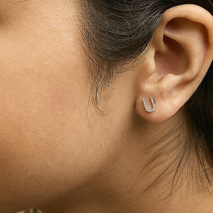 Close-up side view of a model wearing white metal earrings shaped like a small horseshoe, featuring micro-pave set white round gemstones.