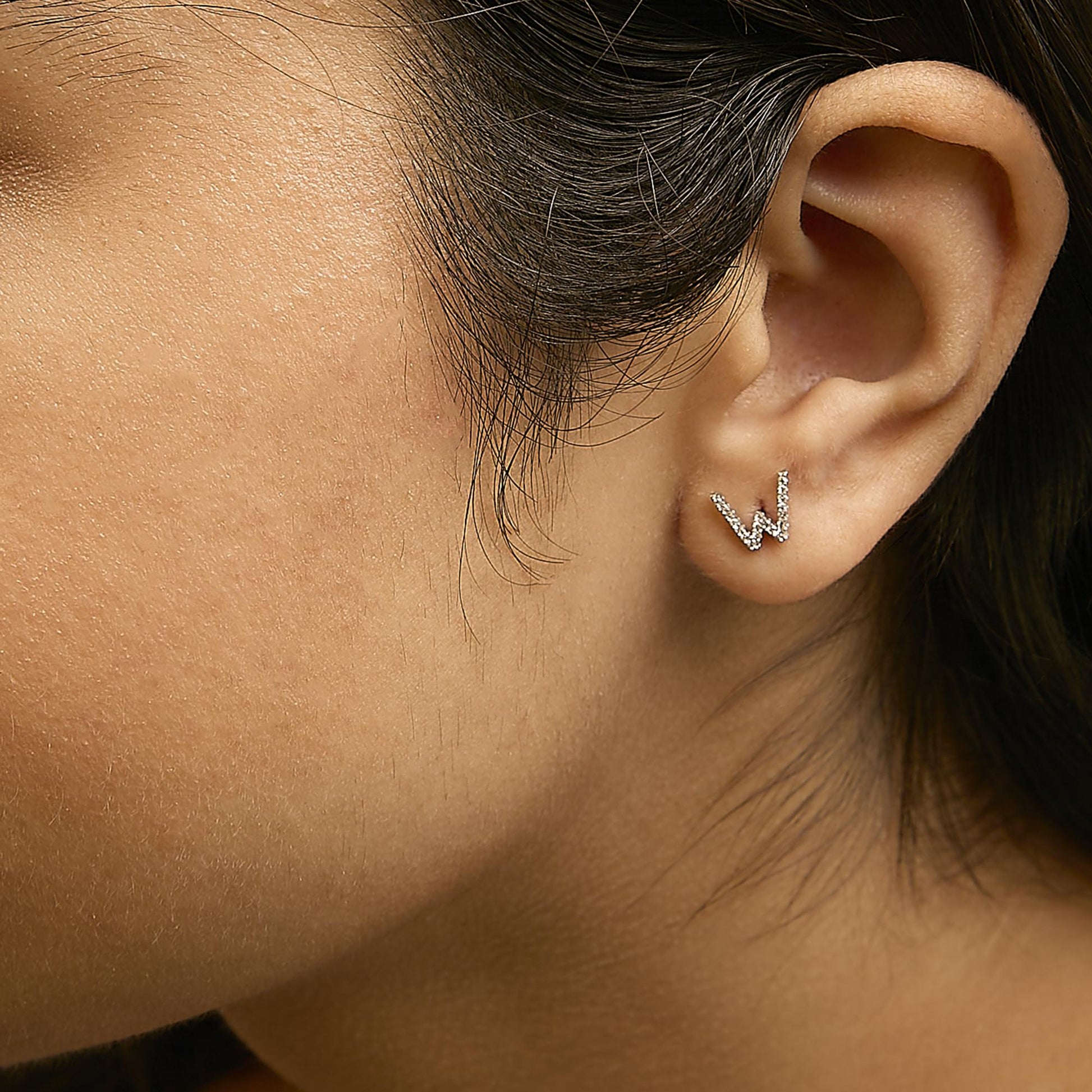 Close-up side view of ear wearing a white metal earring shaped like the letter W, set with small round white gemstones in micro-pave setting.