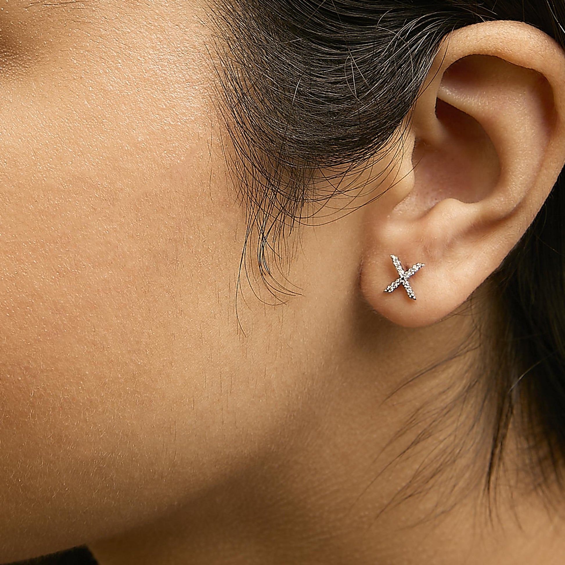 Close-up side view of a model wearing white metal earrings in an X shape, each adorned with small round white gemstones in a micro-pave setting.