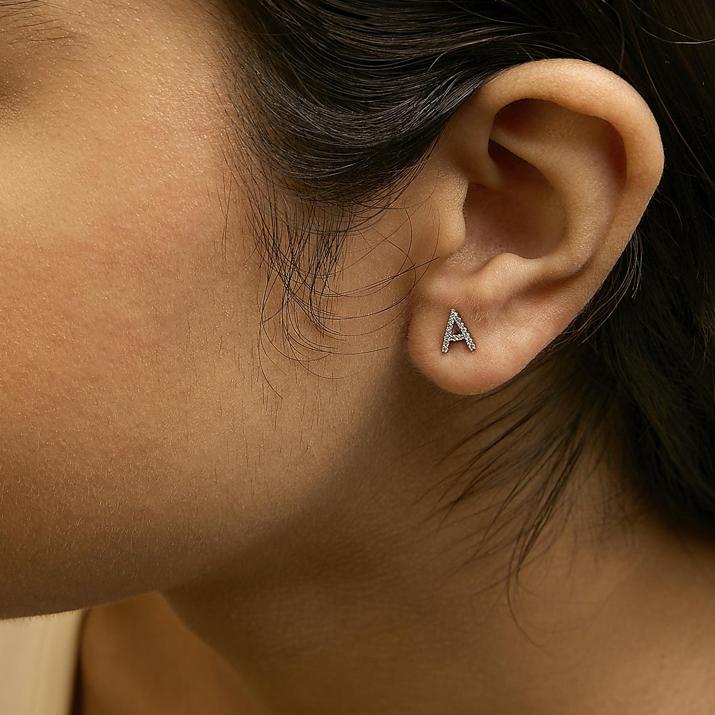 Close-up side view of a model wearing a yellow metal stud earring in the shape of the letter 'A' decorated with white round gemstones in micro-pave setting.