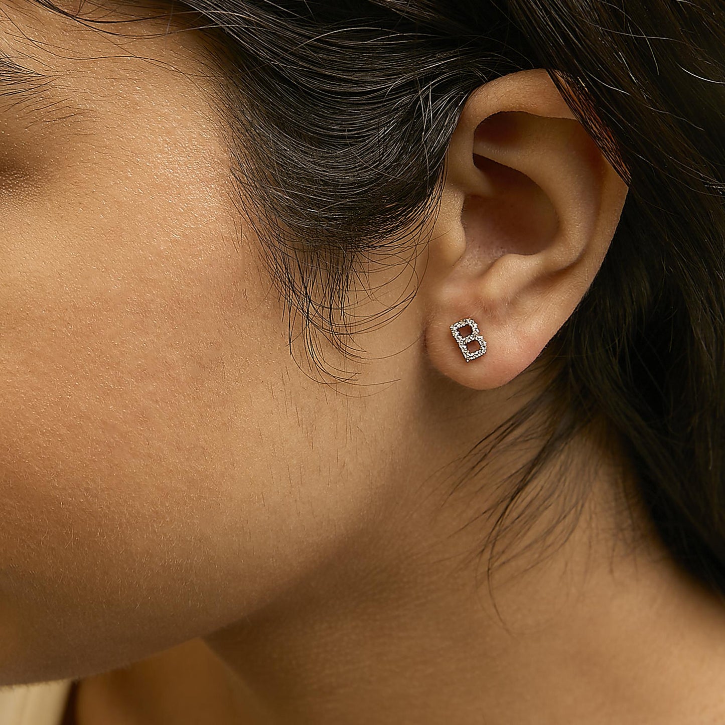 Close-up side view of a model wearing a yellow metal stud earring shaped like the letter B, adorned with small white gemstones in a micro-pave setting.