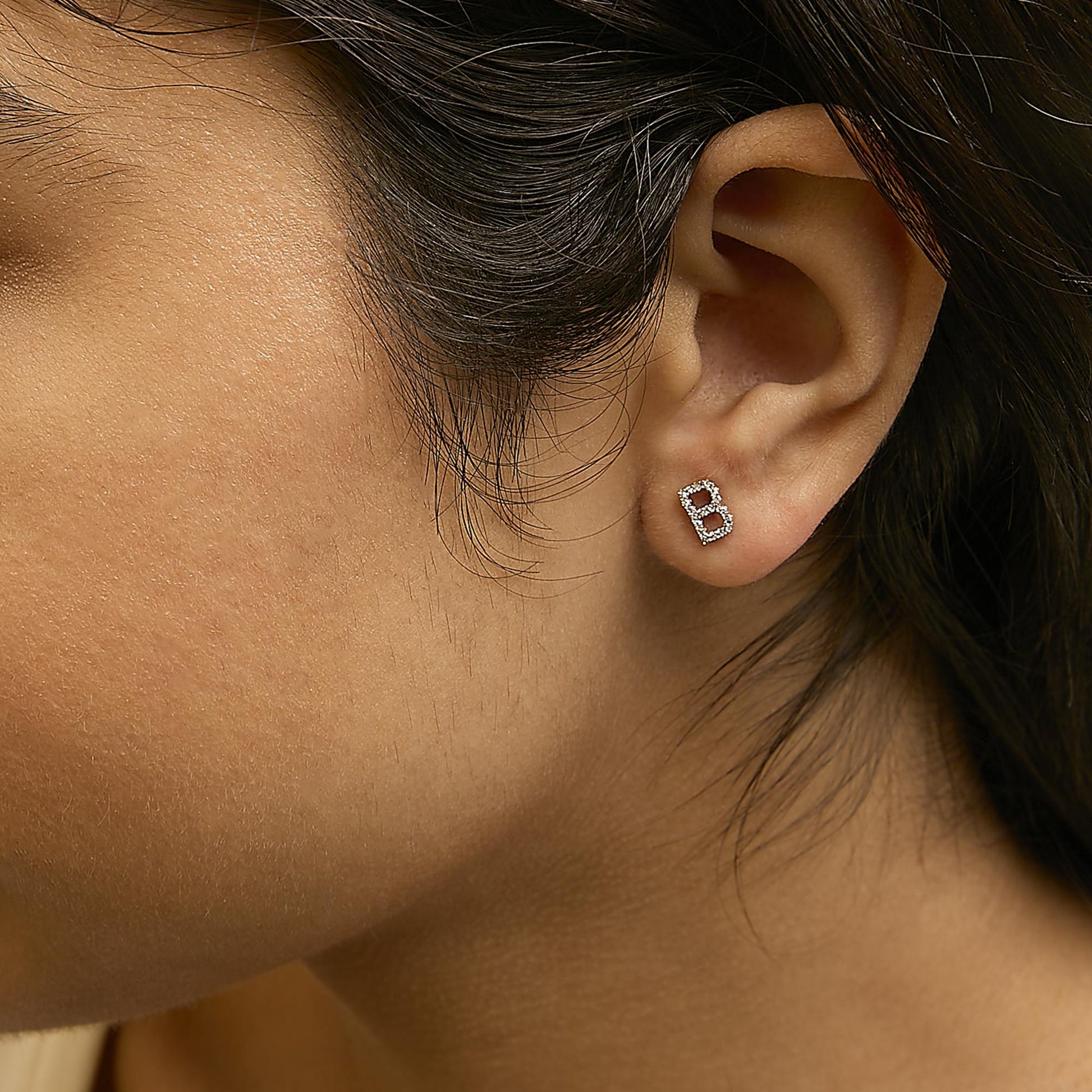 Close-up side view of a model wearing a yellow metal stud earring shaped like the letter B, adorned with small white gemstones in a micro-pave setting.