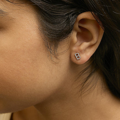 Close-up side view of a model wearing a yellow metal stud earring shaped like the letter B, adorned with small white gemstones in a micro-pave setting.
