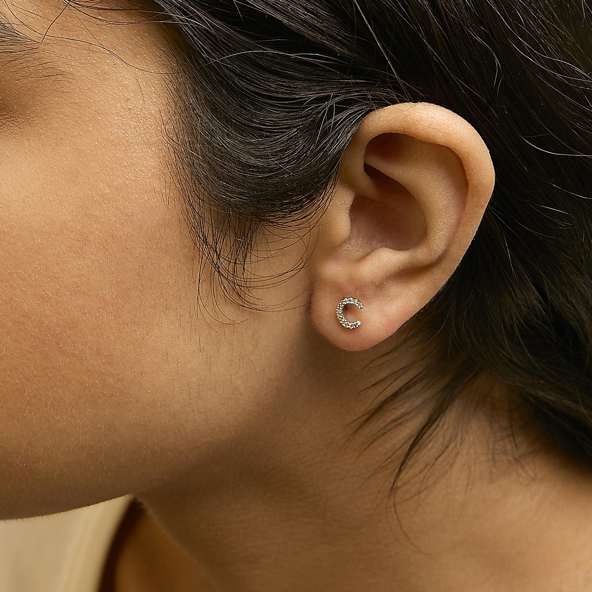 Close-up side angle of a model wearing a small yellow metal earring shaped like the letter C, adorned with micro-pave set white round gemstones.