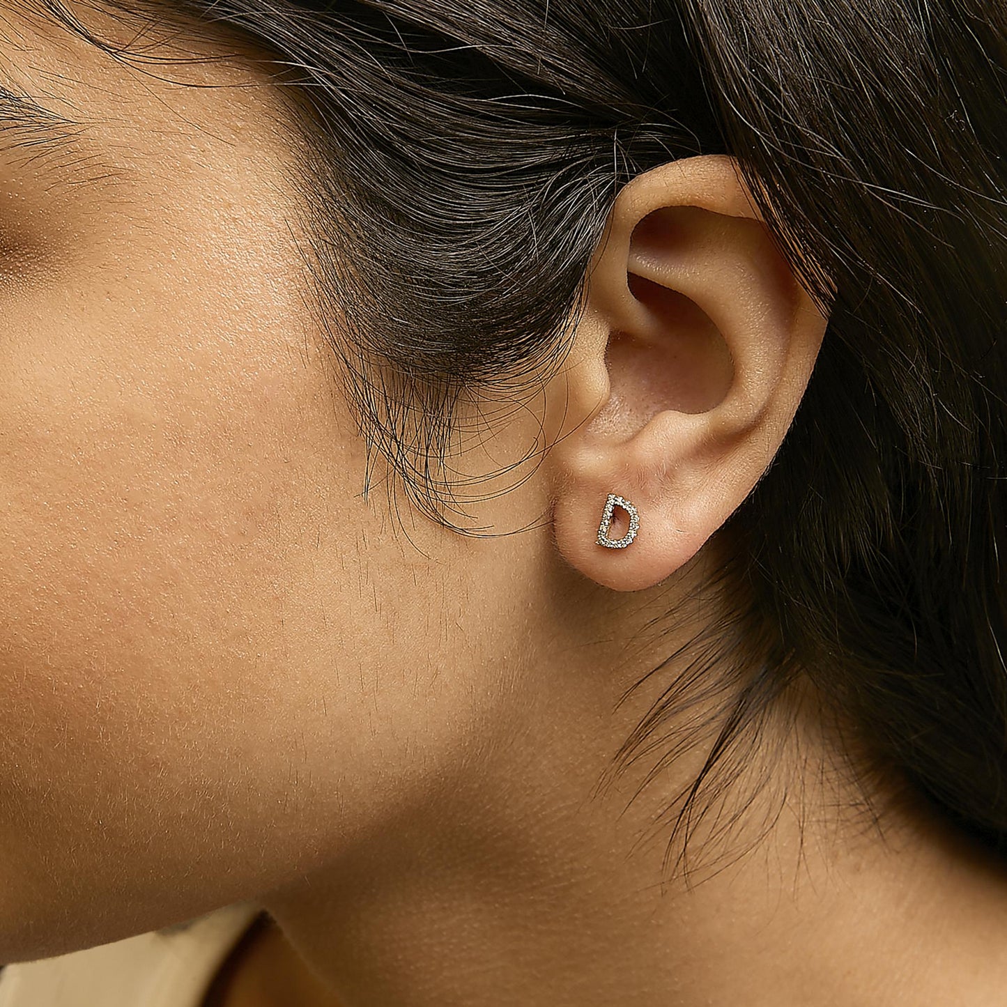 Close-up side view of a model wearing yellow metal stud earrings shaped like the letter D, set with small white gemstones in a micro-pave setting.