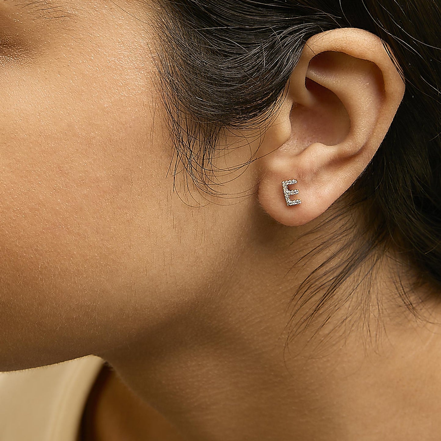 Close-up side view of a model wearing a yellow metal earring shaped like the letter 'E' with white round micro-pave gemstones.