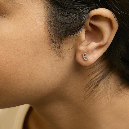 Close-up side view of a model wearing a yellow metal earring shaped like the letter 'E' with white round micro-pave gemstones.