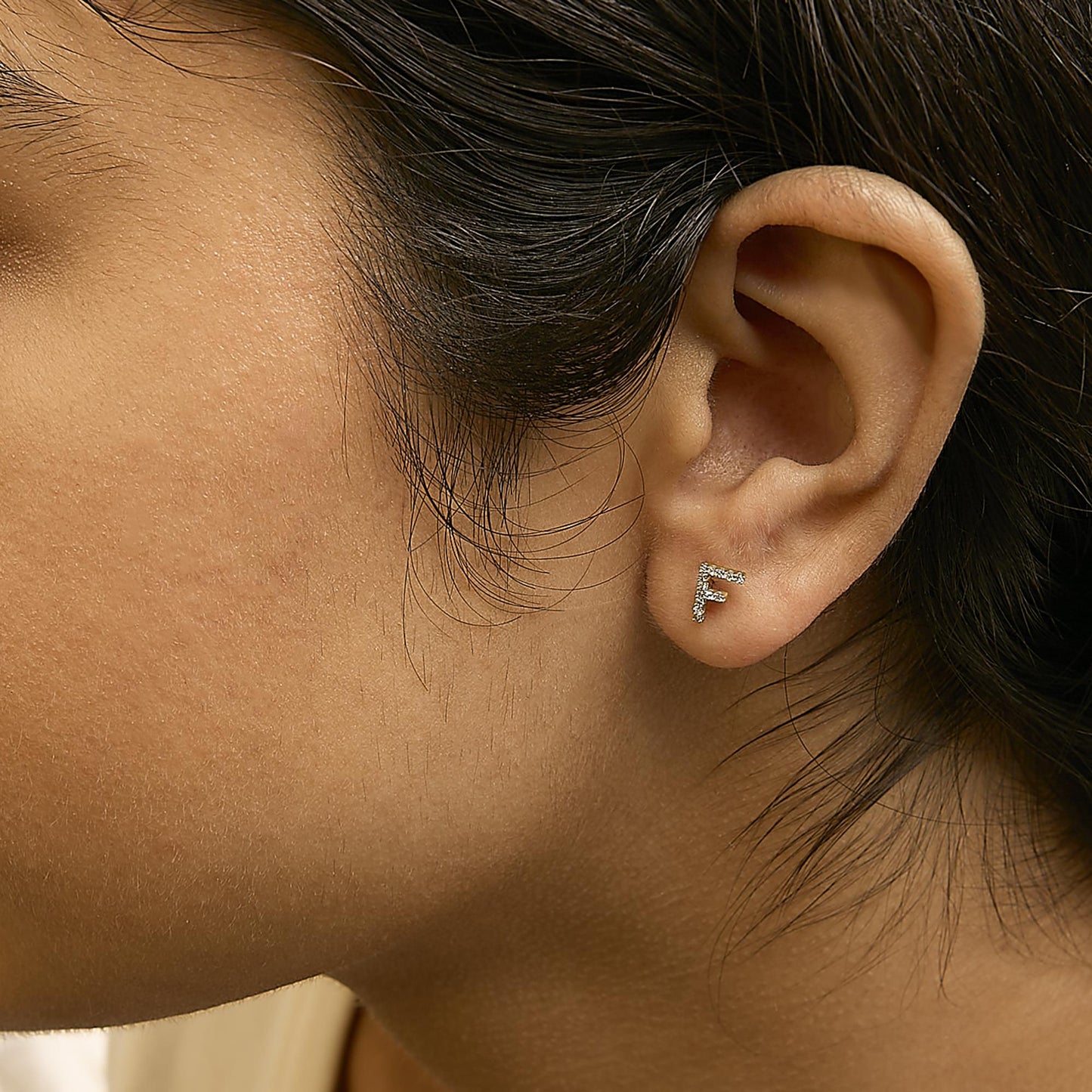 Close-up side view of a model wearing a yellow metal earring shaped like the letter F, adorned with small white gemstones in a micro-pave setting.