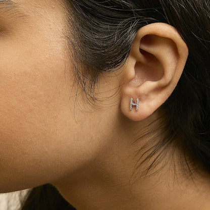 Close-up side view of a model's ear wearing a yellow metal earring shaped like the letter 'H' adorned with small white gemstones in a micro-pave setting.