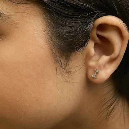 Close-up side view of a model wearing a small yellow metal earring shaped like a horizontal bar with white round gemstones in micro-pave setting.