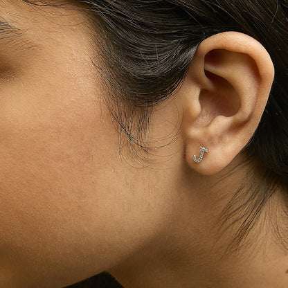 Close-up side view of a model wearing a small yellow metal earring shaped like the letter J, adorned with multiple round white gemstones in a micro-pave setting.