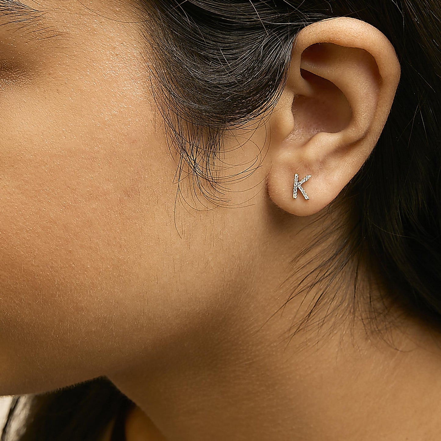 Close-up side view of a model wearing a yellow metal earring shaped like the letter K, adorned with small white gemstones in a micro-pave setting.