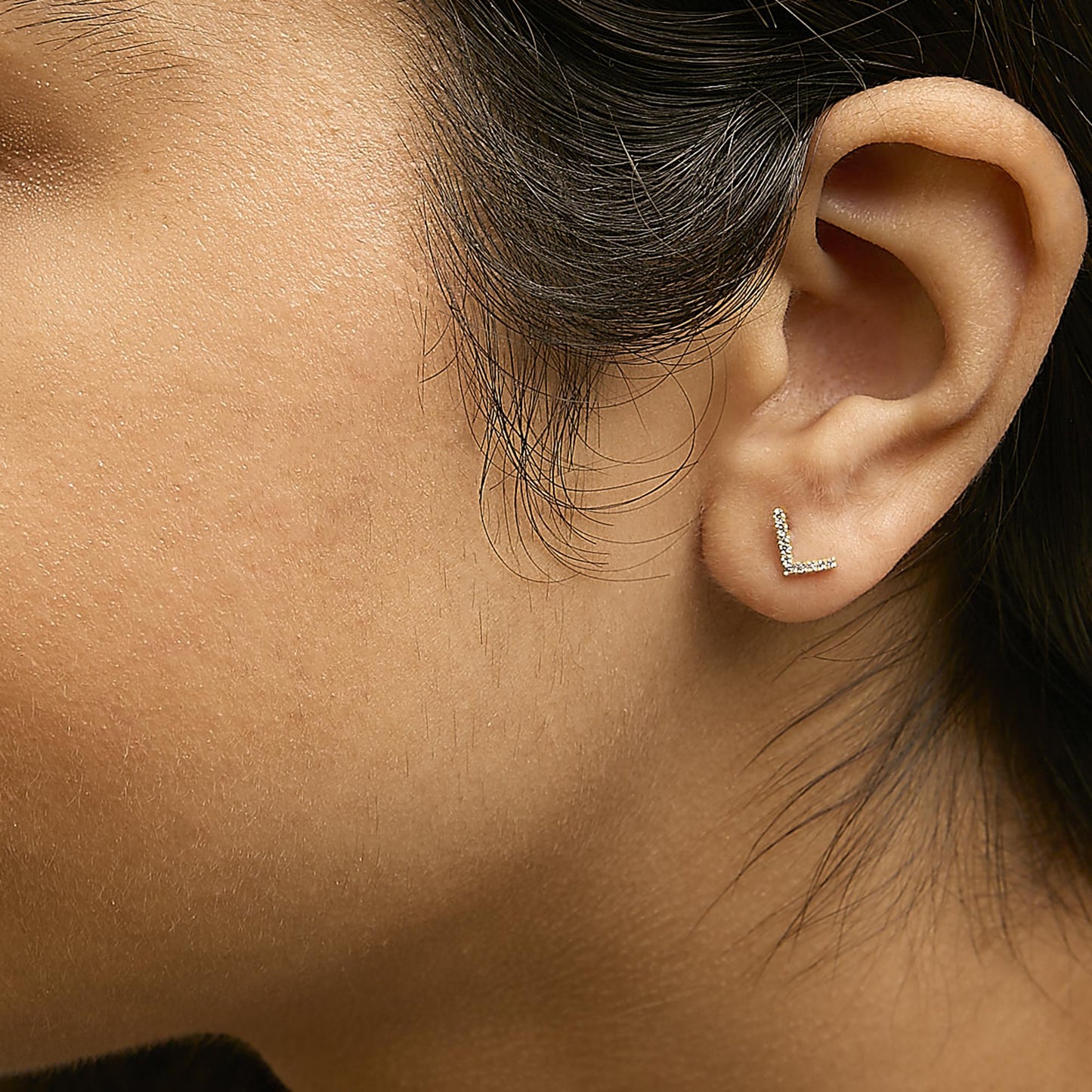 Close-up side view of a model wearing yellow metal earrings in an L-shape with white round gemstones set in a micro-pave style.