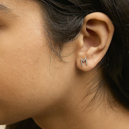 Close-up side angle of a model wearing a yellow metal stud earring shaped like the letter M, decorated with multiple small white gemstones in a micro-pave setting.