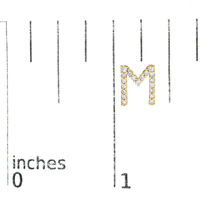 Close-up of a yellow metal earring shaped like the letter M, adorned with small round white gemstones in a micro-pave setting, shown on a scale with inch measurements.