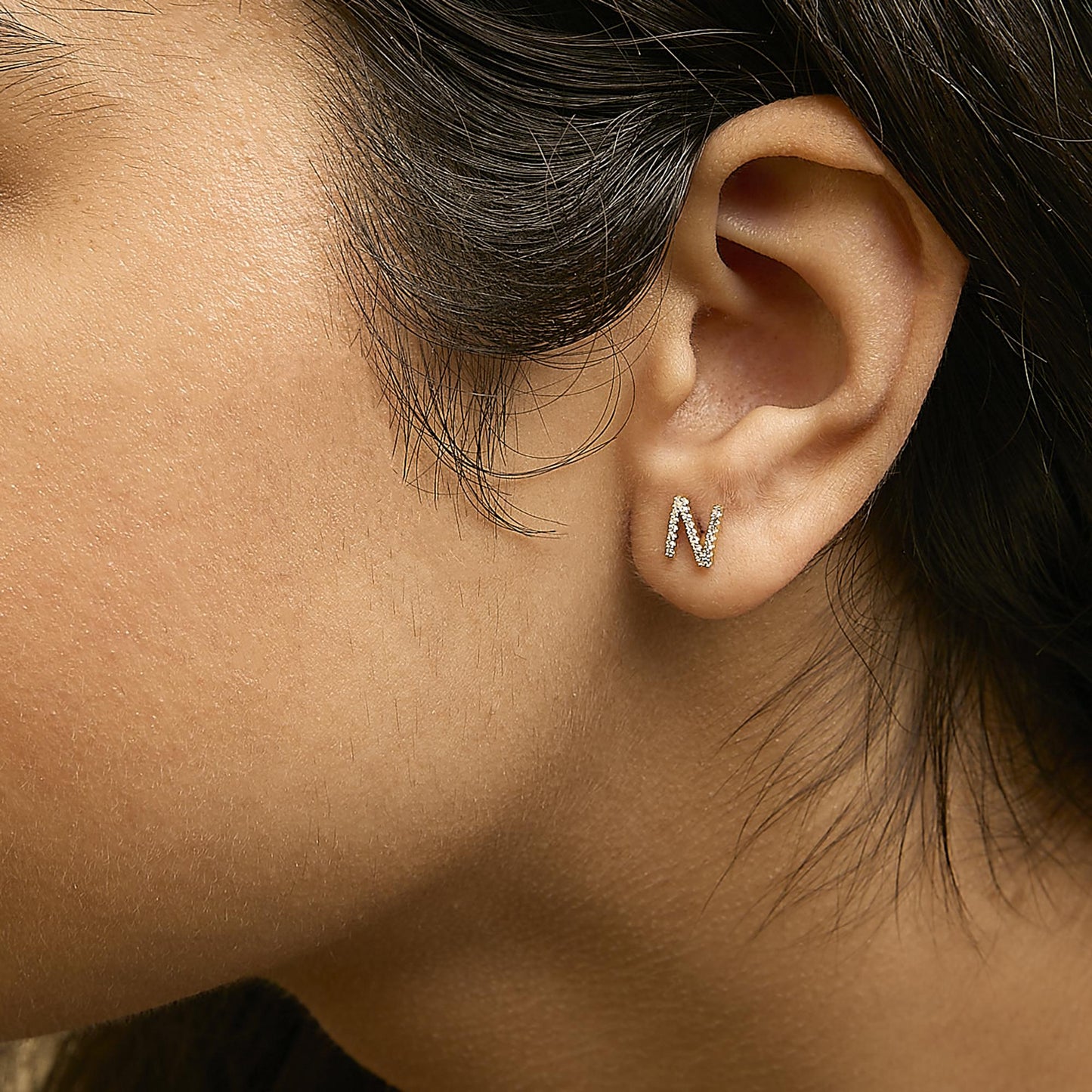 Close-up side view of a model wearing a small yellow metal earring shaped like the letter 'N,' accented with white gemstones in a micro-pave setting.