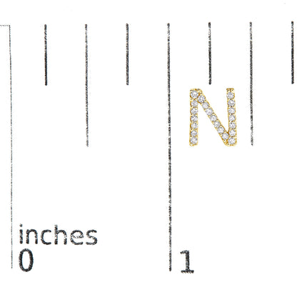 Close-up of a yellow metal earring shaped like the letter N, adorned with small white gemstones, shown on a scale with inch markings, photographed from the front.