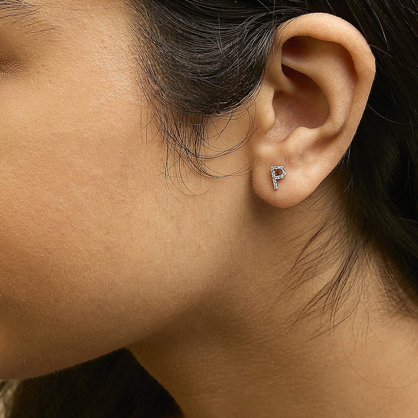 Close-up side view of a model wearing a yellow metal earring shaped like the letter P, adorned with small white gemstones in a micro-pave setting.