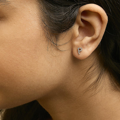 Close-up side view of a model wearing a yellow metal earring shaped like the letter P, adorned with small white gemstones in a micro-pave setting.