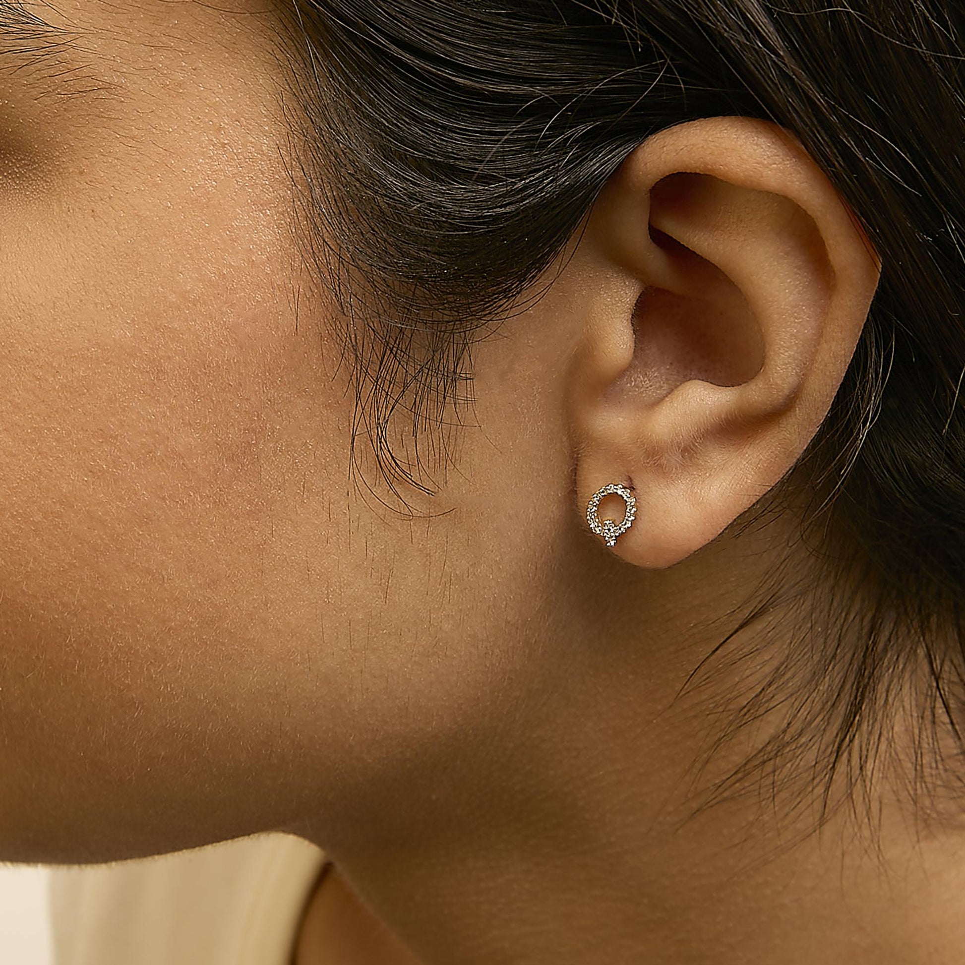 Close-up side view of a model wearing a pair of small yellow metal circular stud earrings with white round gemstones in a micro-pave setting.