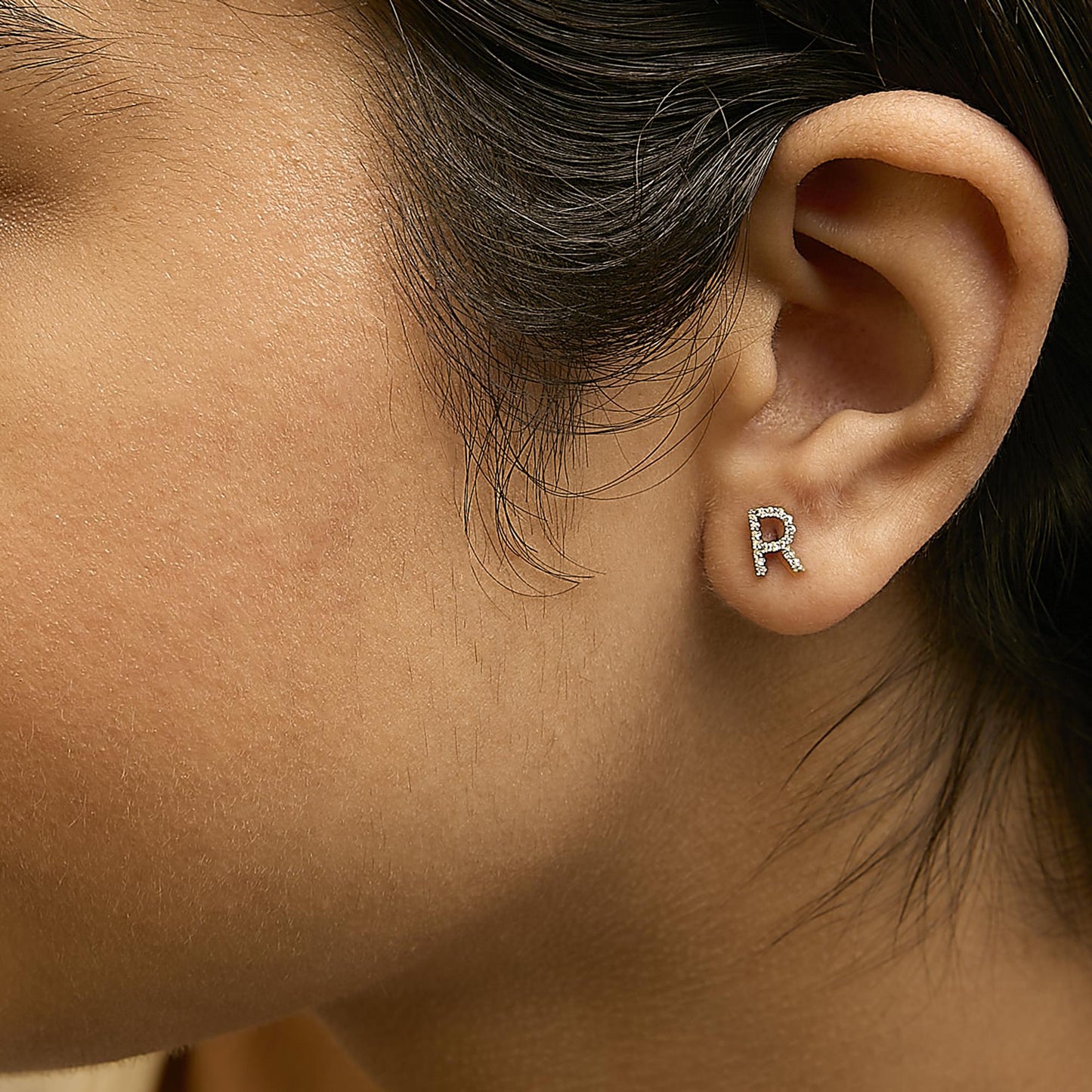 Close-up side view of a model wearing a yellow metal earring shaped like the letter 'R' with round white gemstones in a micro-pave setting.