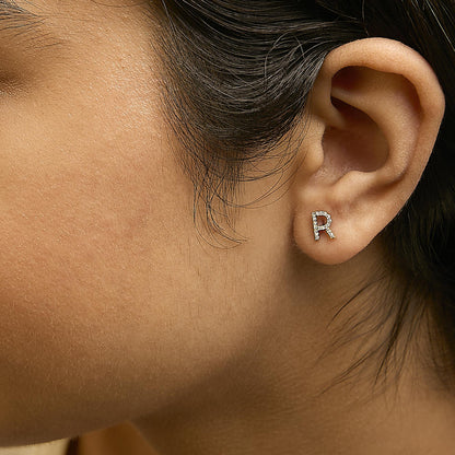 Close-up side view of a model wearing a yellow metal earring shaped like the letter 'R' with round white gemstones in a micro-pave setting.