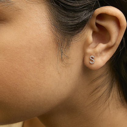Close-up side view of a model wearing yellow metal earrings shaped like the letter S, adorned with small white gemstones in a micro-pave setting.
