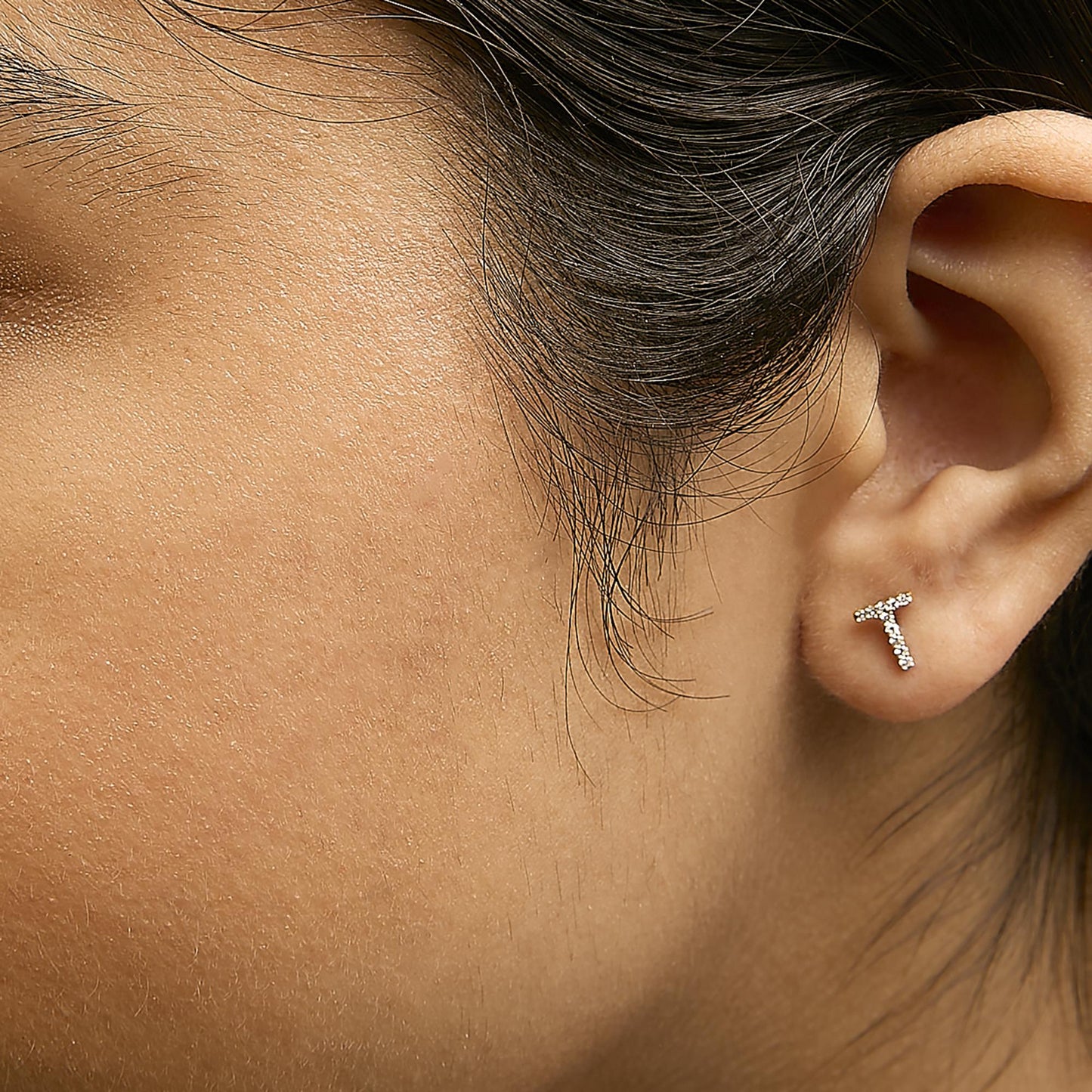 Close-up side angle of a model wearing yellow metal earrings shaped like the letter T, studded with small white gemstones in a micro-pave setting.