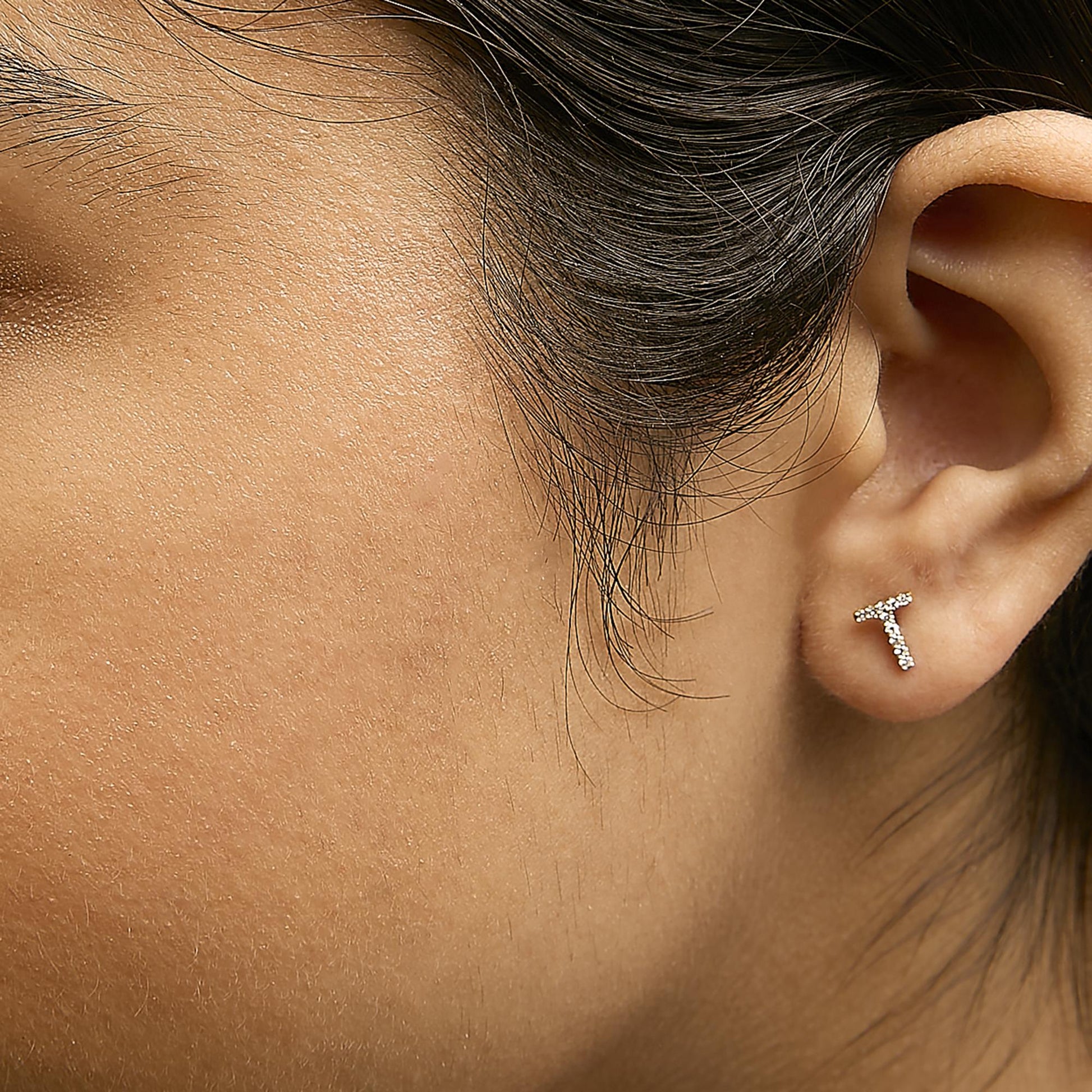 Close-up side angle of a model wearing yellow metal earrings shaped like the letter T, studded with small white gemstones in a micro-pave setting.