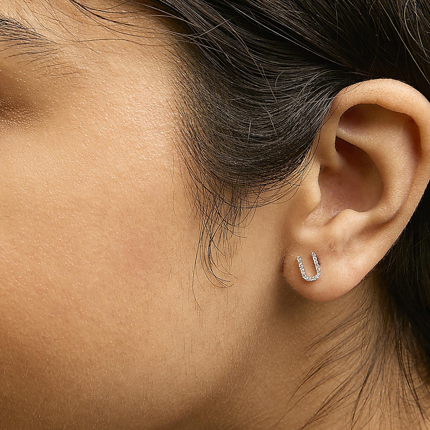 Close-up side view of a model wearing yellow metal earrings shaped like an open U, decorated with small white gemstones in a micro-pave setting.