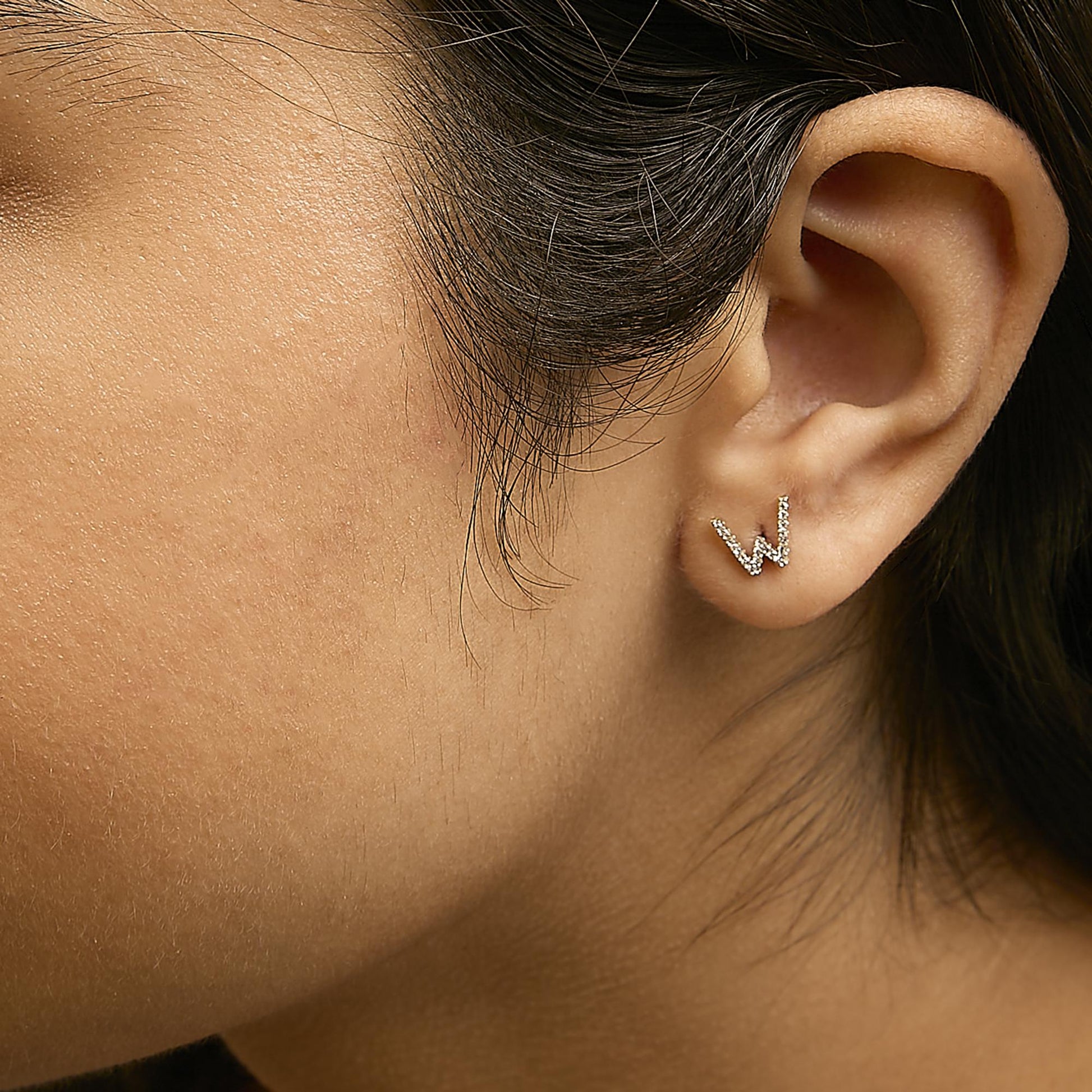 Close-up side view of a model wearing yellow metal earrings shaped like the letter W, adorned with small white gemstones in a micro-pave setting.