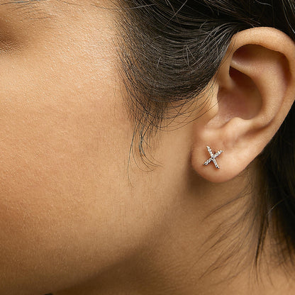 Close-up side view of a model wearing a pair of yellow metal earrings shaped like an 'X' studded with small white gemstones in a micro-pave setting.