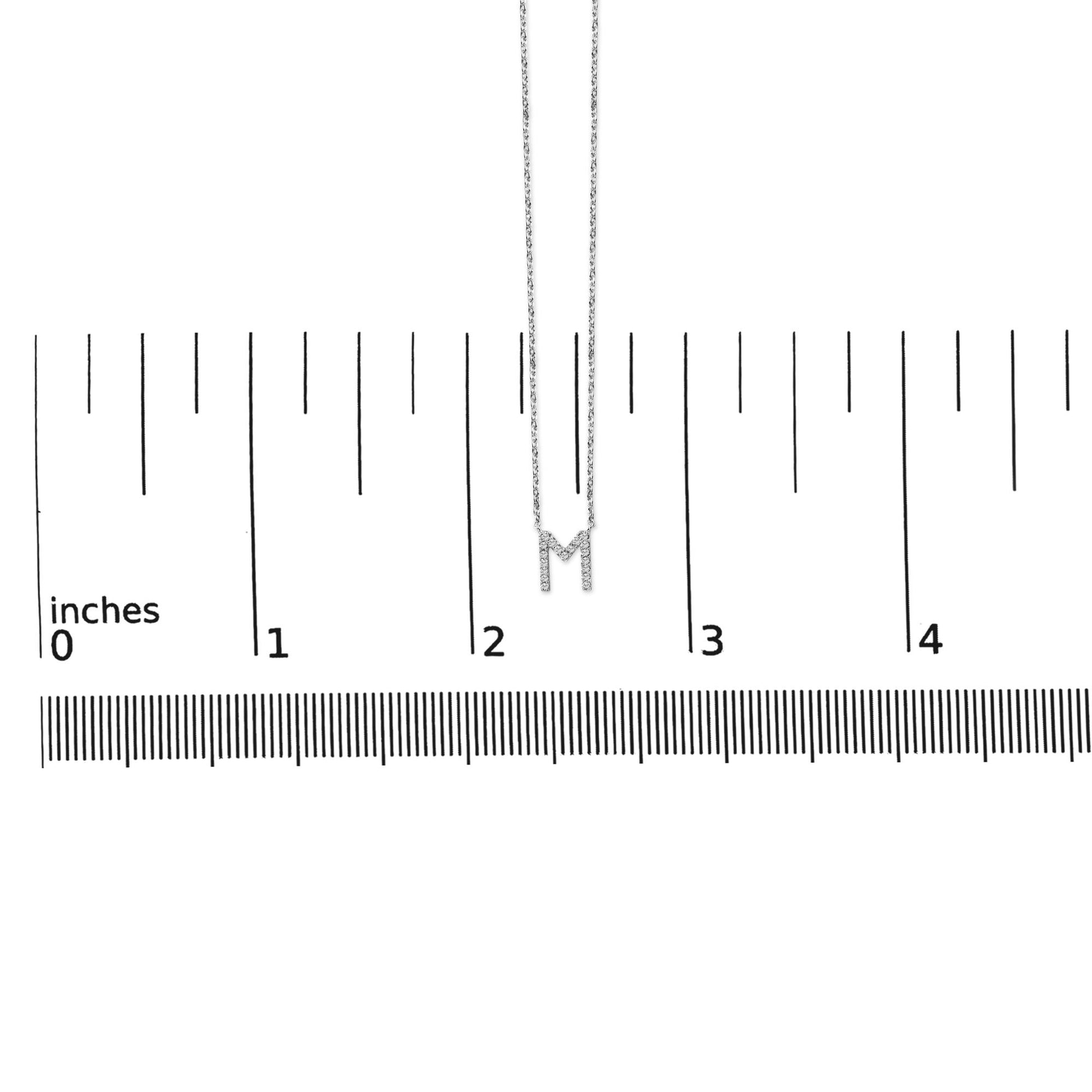 Close-up front view of a white metal necklace with a letter 'M' pendant accented with white round gemstones in a micro-pave setting, shown on a measurement scale.