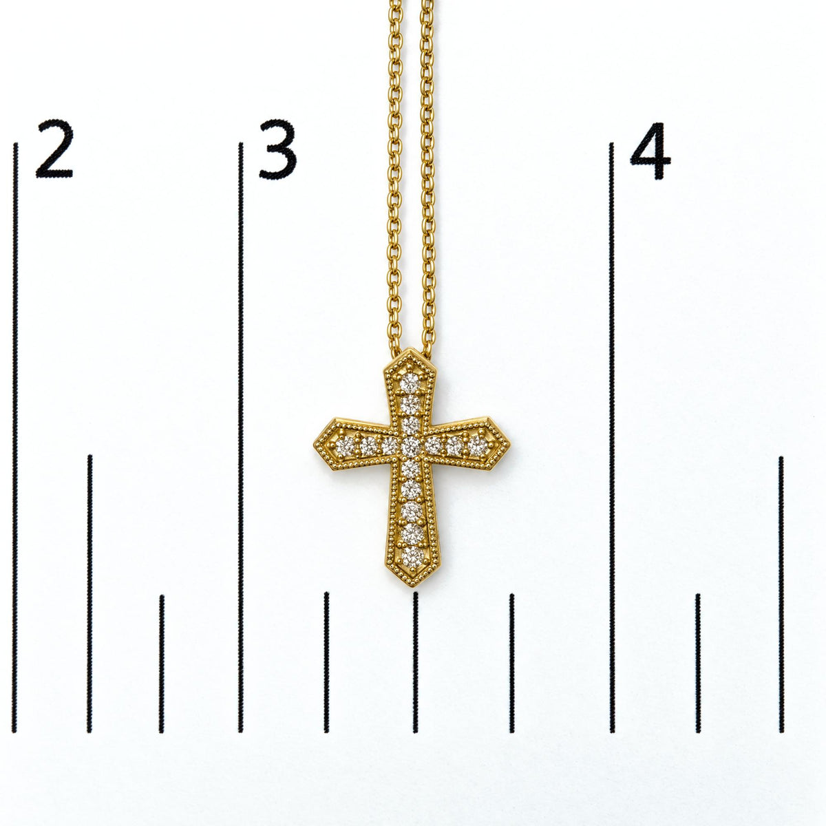 Close-up front view of a yellow metal cross-shaped necklace pendant with round white gemstones in pave setting, shown on a scale.