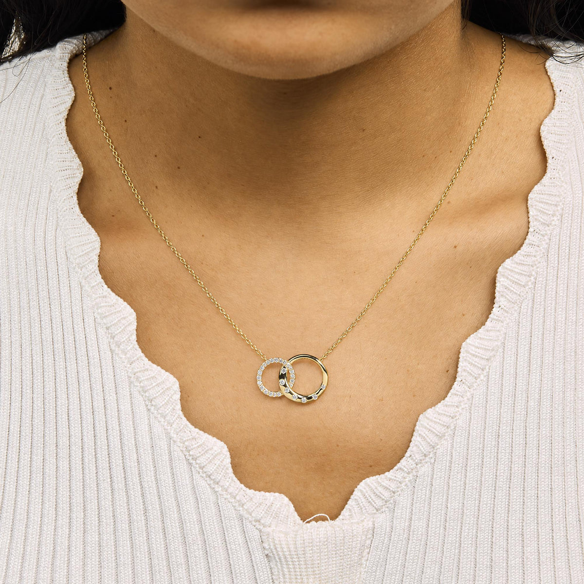 Close-up front view of a model wearing a yellow metal necklace with two interlocking circular pendants, one adorned with small white gemstones in a prong setting and the other with scattered white gemstones.