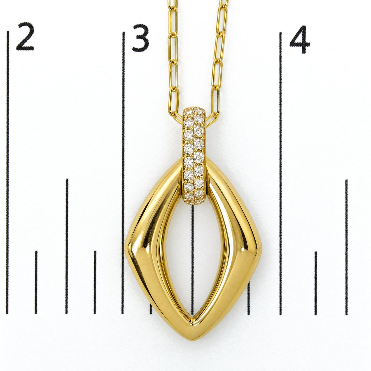 Close-up front view of yellow metal necklace pendant with an elongated diamond-shaped design and a row of round white gemstones set in a prong setting on the bail, shown on a measurement scale.